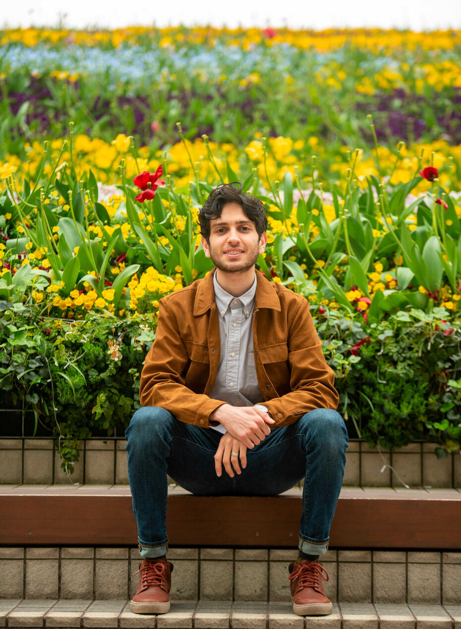 a man sitting on a wooden bench, and behind him there is a lot of greenery and flowers.